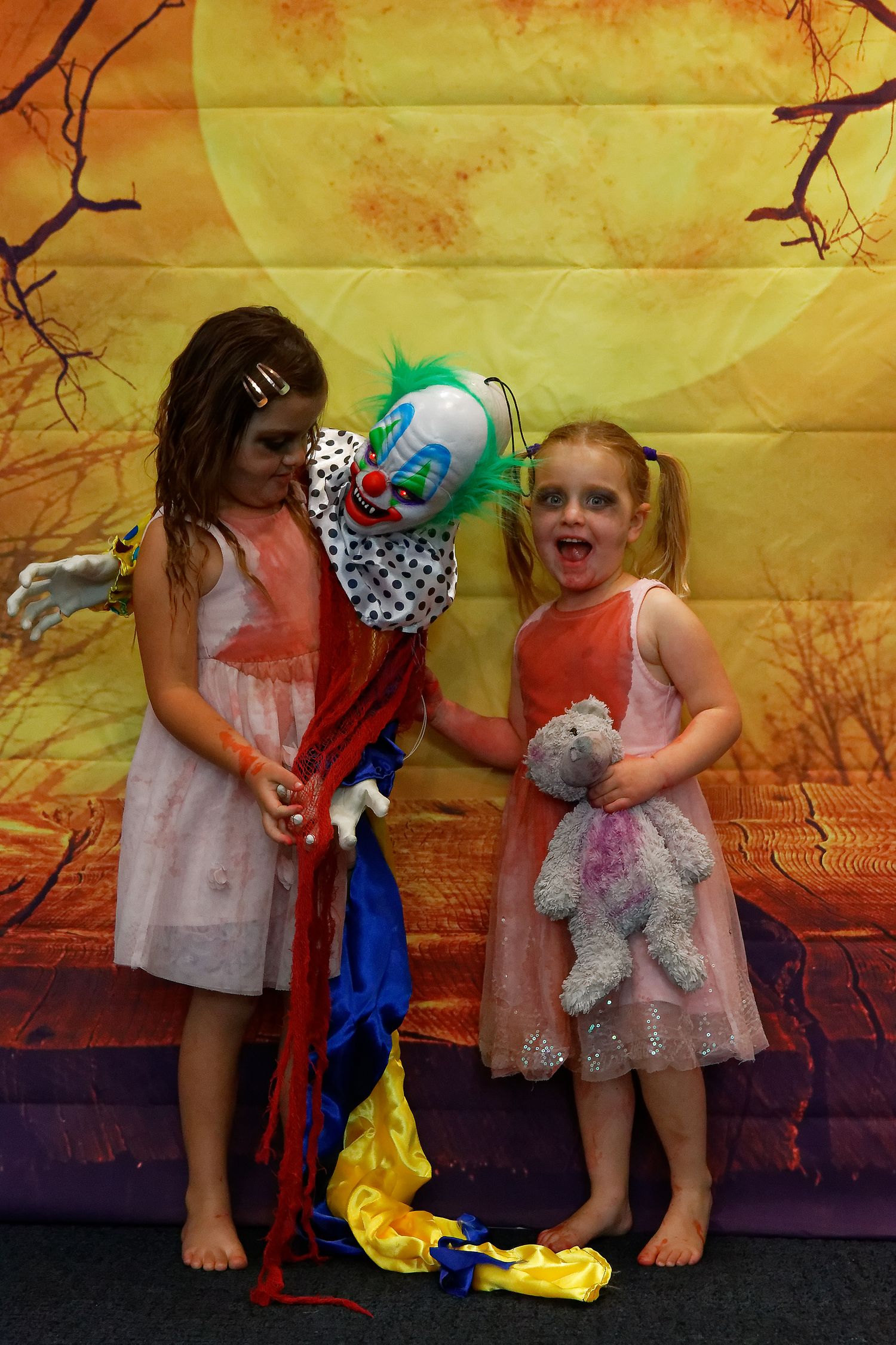 Two children in dresses posing with colorful clown prop against orange halloween backdrop.