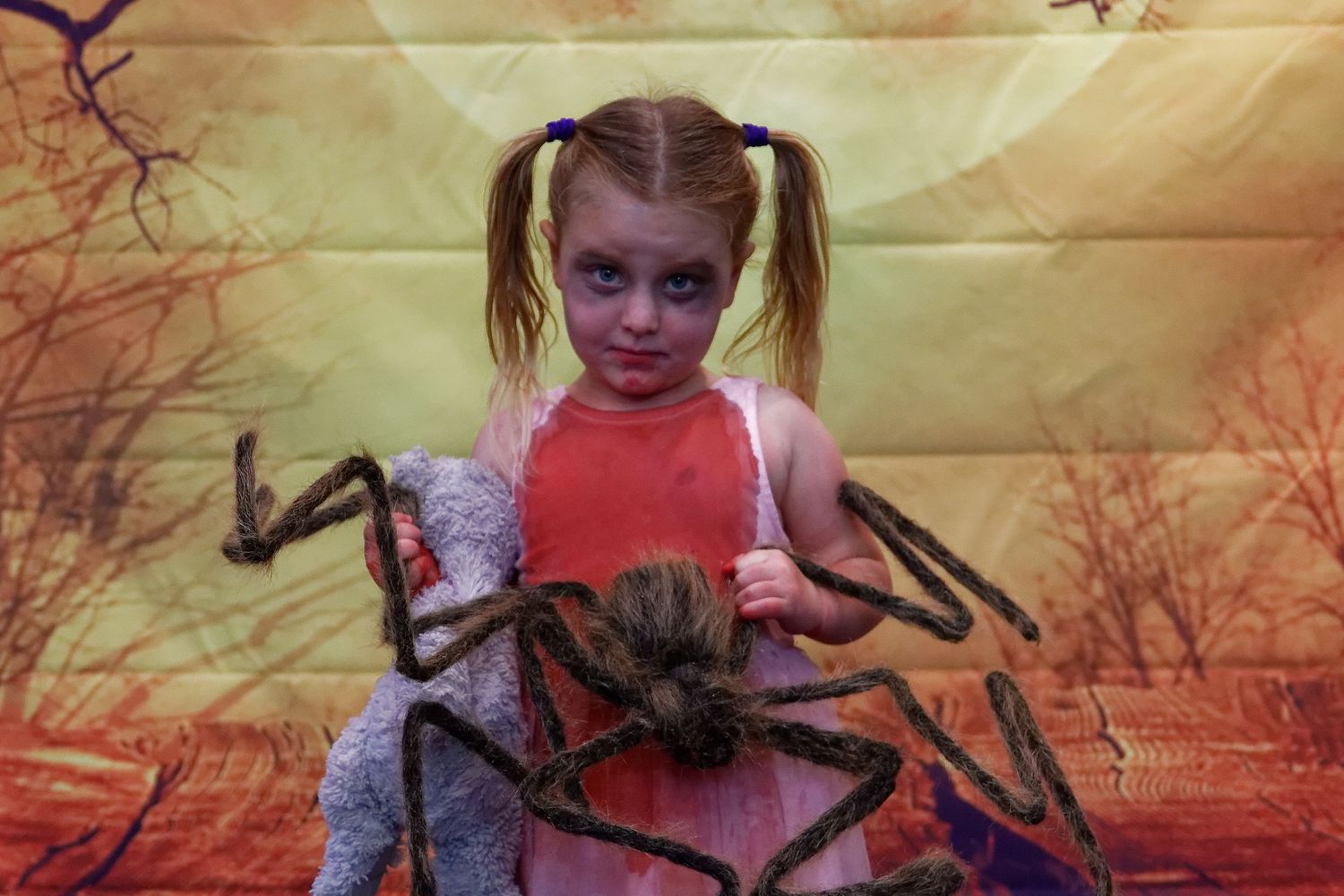 Child with pigtails poses with large spider prop against orange sunset background.