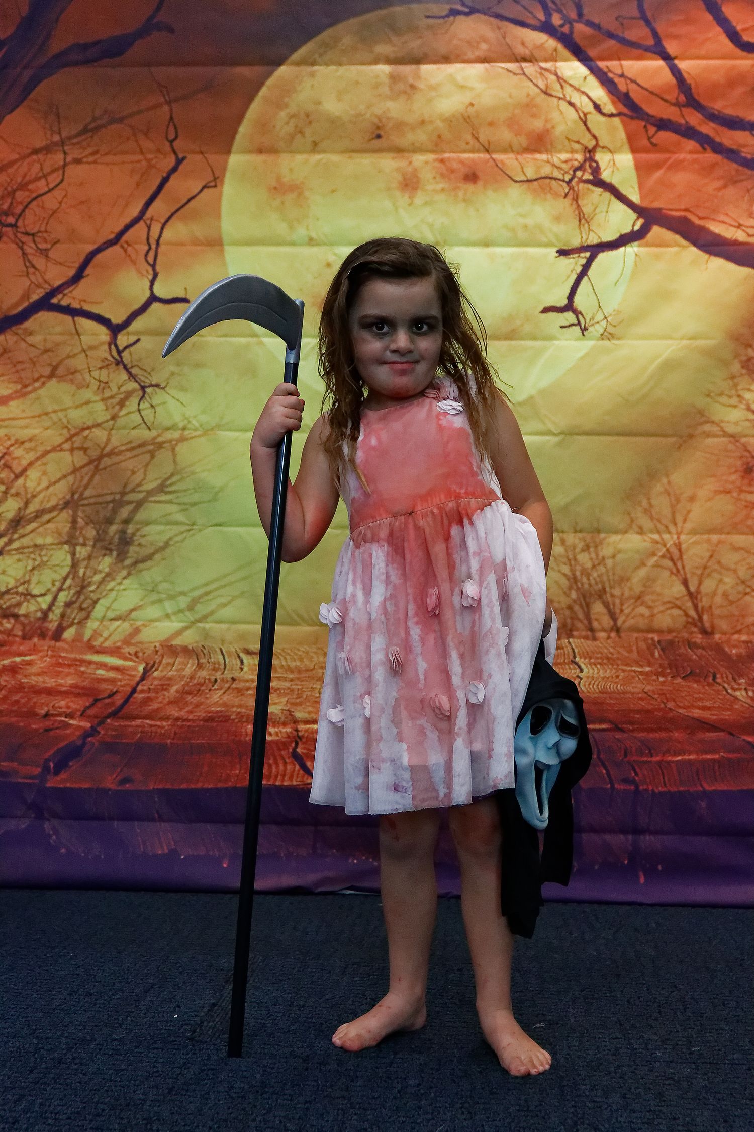 Child in blood-stained dress holds scythe prop against spooky orange backdrop.