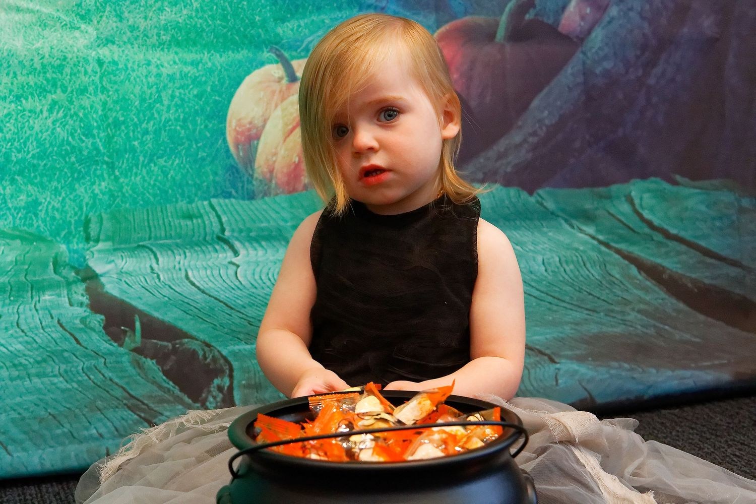 Child in black dress sits with halloween cauldron prop against green backdrop.