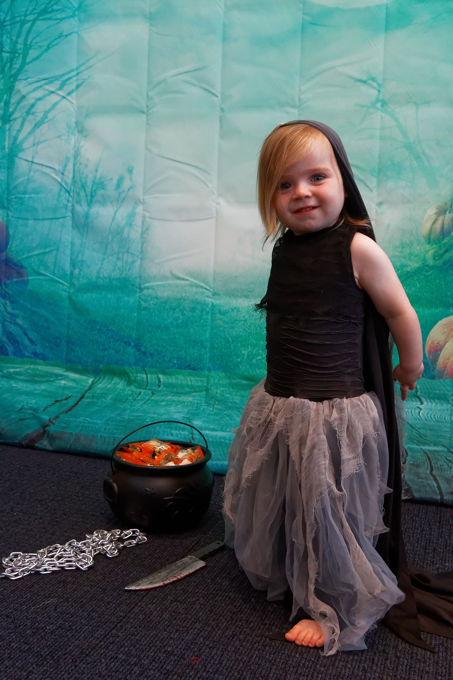 Child in Halloween costume with gray tulle skirt standing next to decorative cauldron prop.