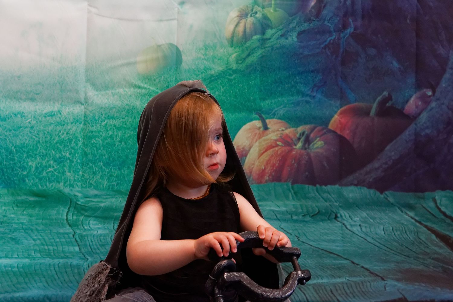 Child in black outfit examining props against turquoise backdrop with pumpkins.