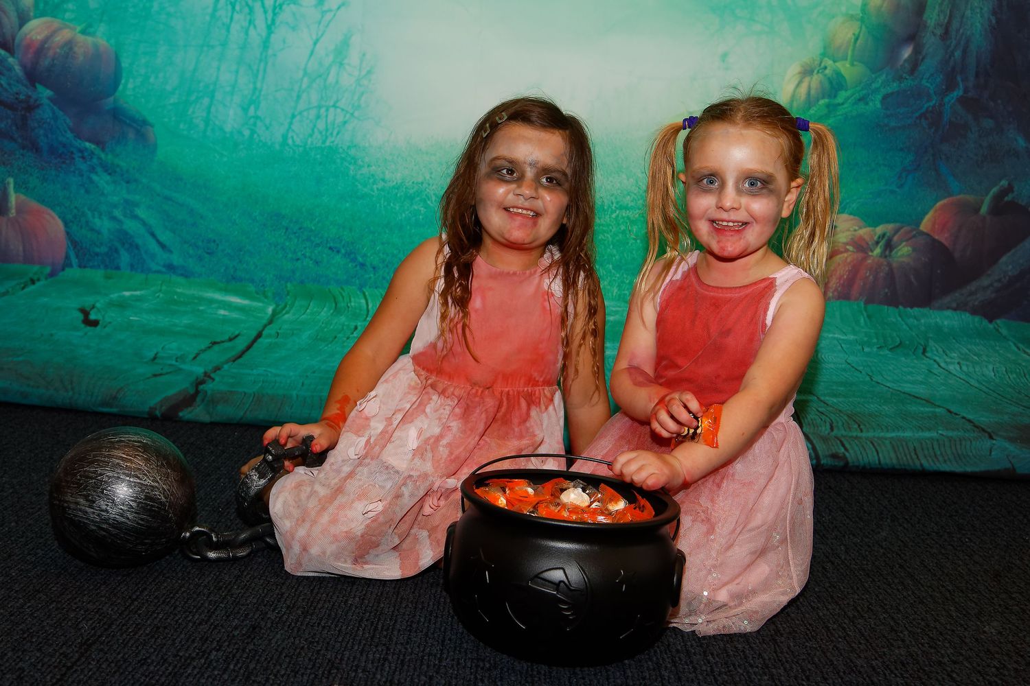 Two children in pink dresses sit with Halloween cauldrons against a spooky green backdrop.
