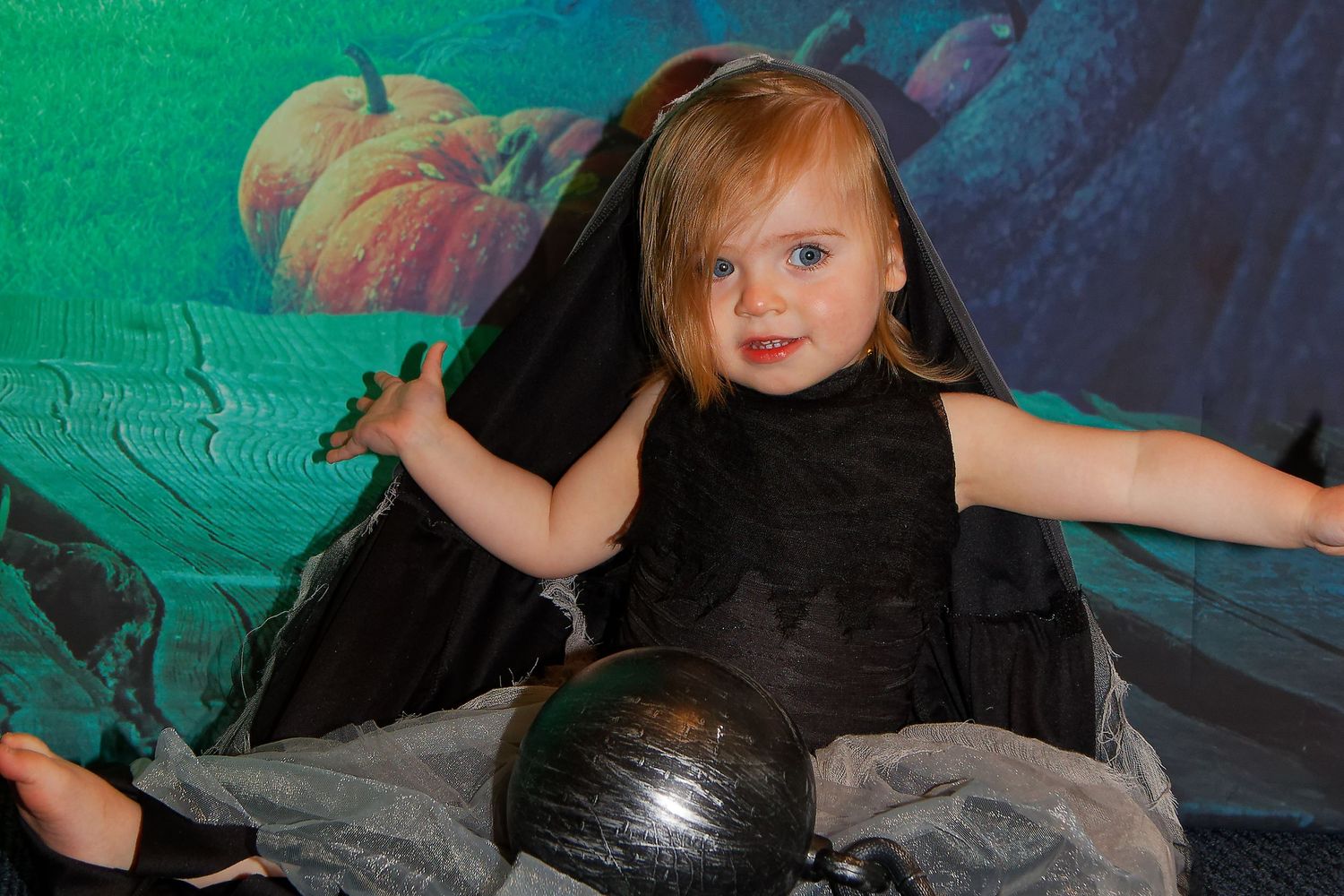 Child in black dress poses with Halloween props against spooky forest backdrop.