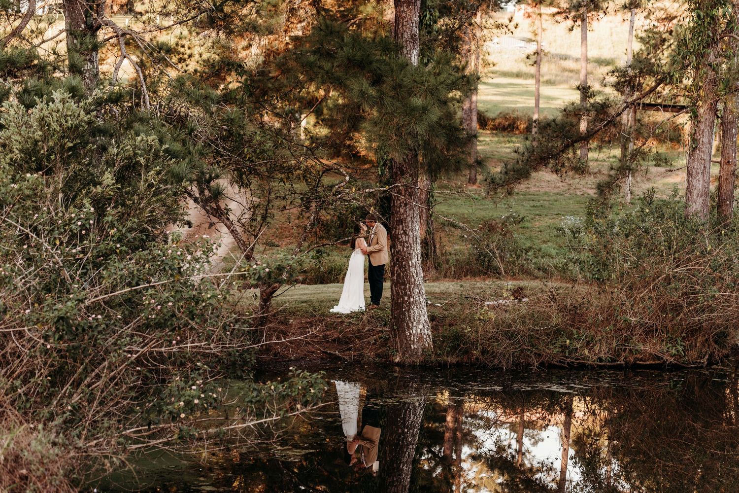 Wedding couple's reflection in a serene pond surrounded by autumn trees.