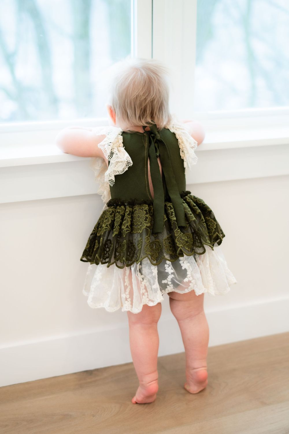 A toddler in a frilly green and white dress stands by a window, looking out, with her back to the camera.