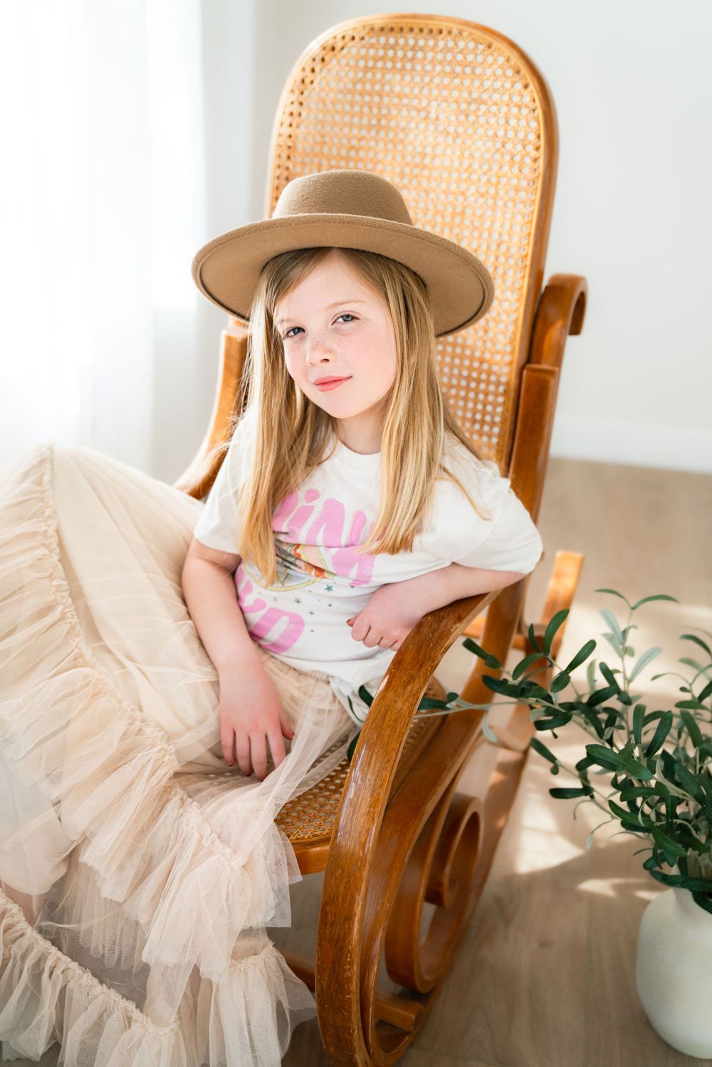 A young girl wearing a stylish beige hat and a white t-shirt paired with a tulle skirt sitting in a wooden rocking chair