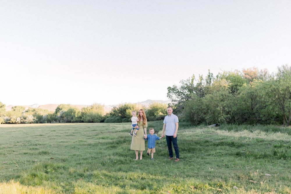 Family of four standing in green meadow with mountains and trees in golden hour light.