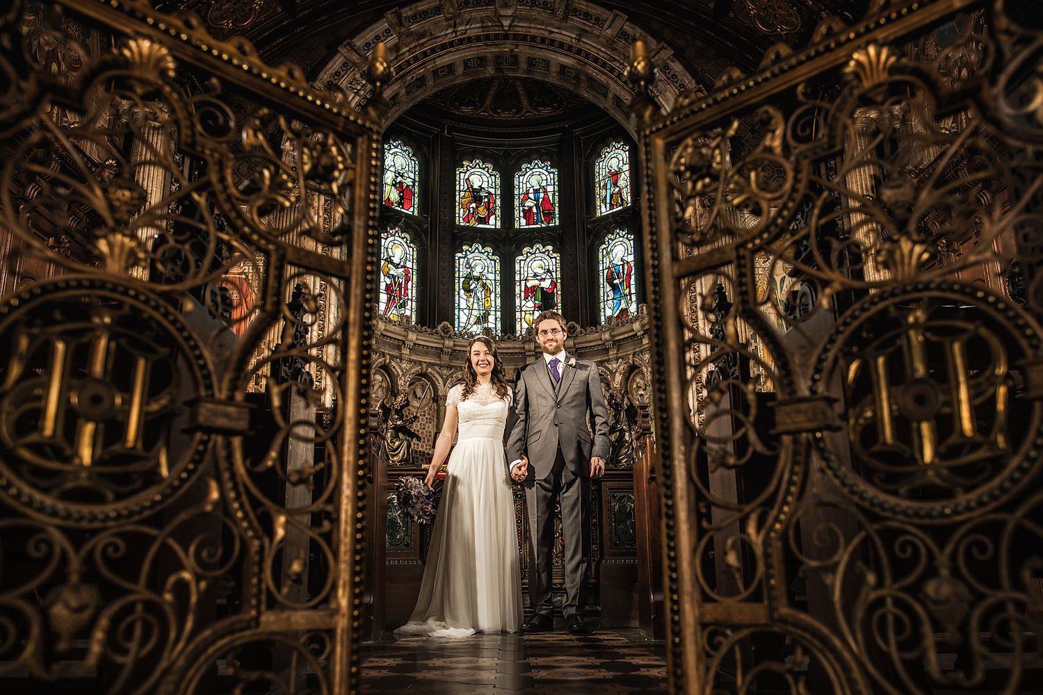 Wedding couple stands in gothic chapel with ornate carved details and stained glass windows.