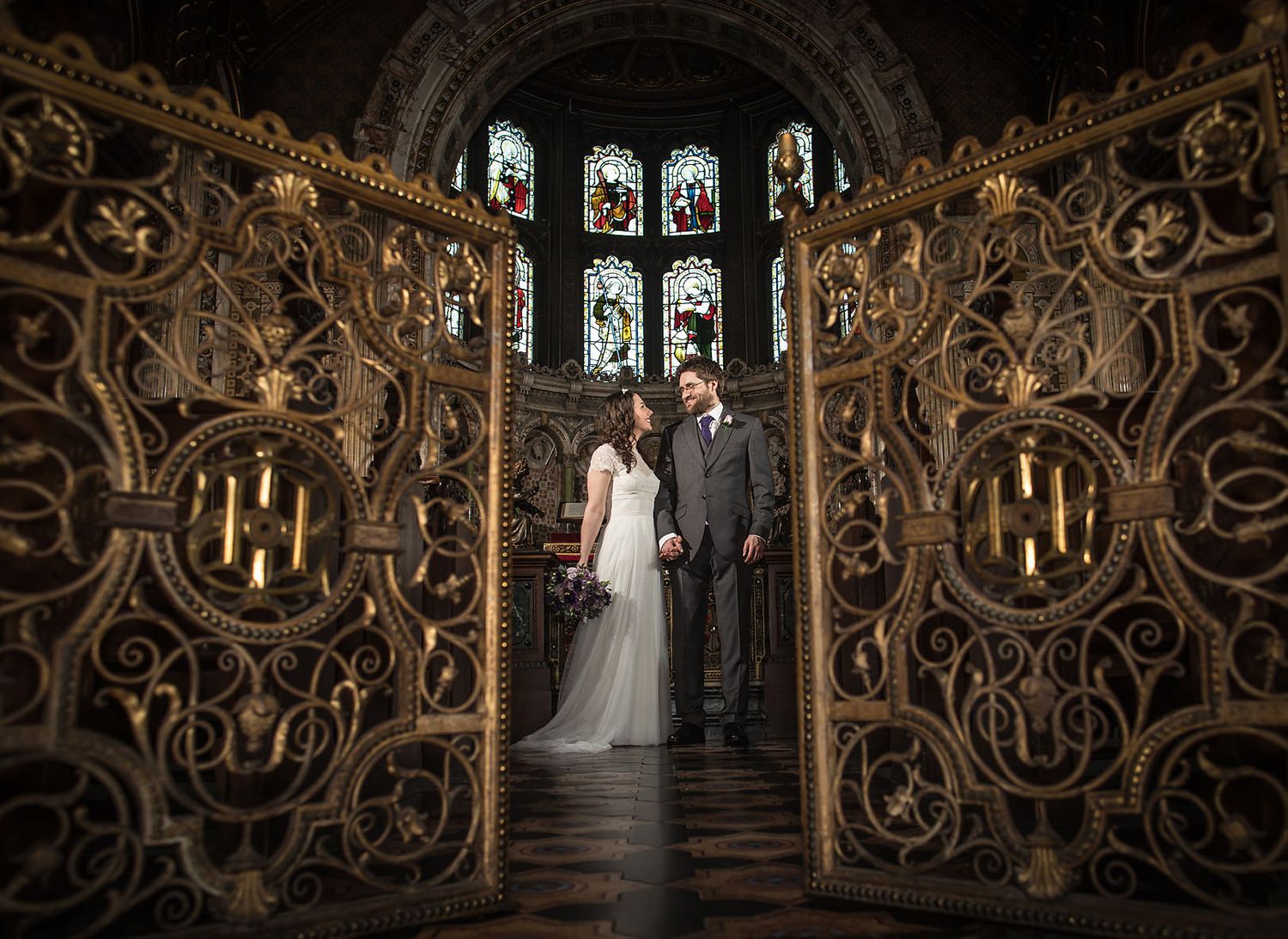 Couple framed by elaborate carved wooden archway with stained glass windows in gothic setting.