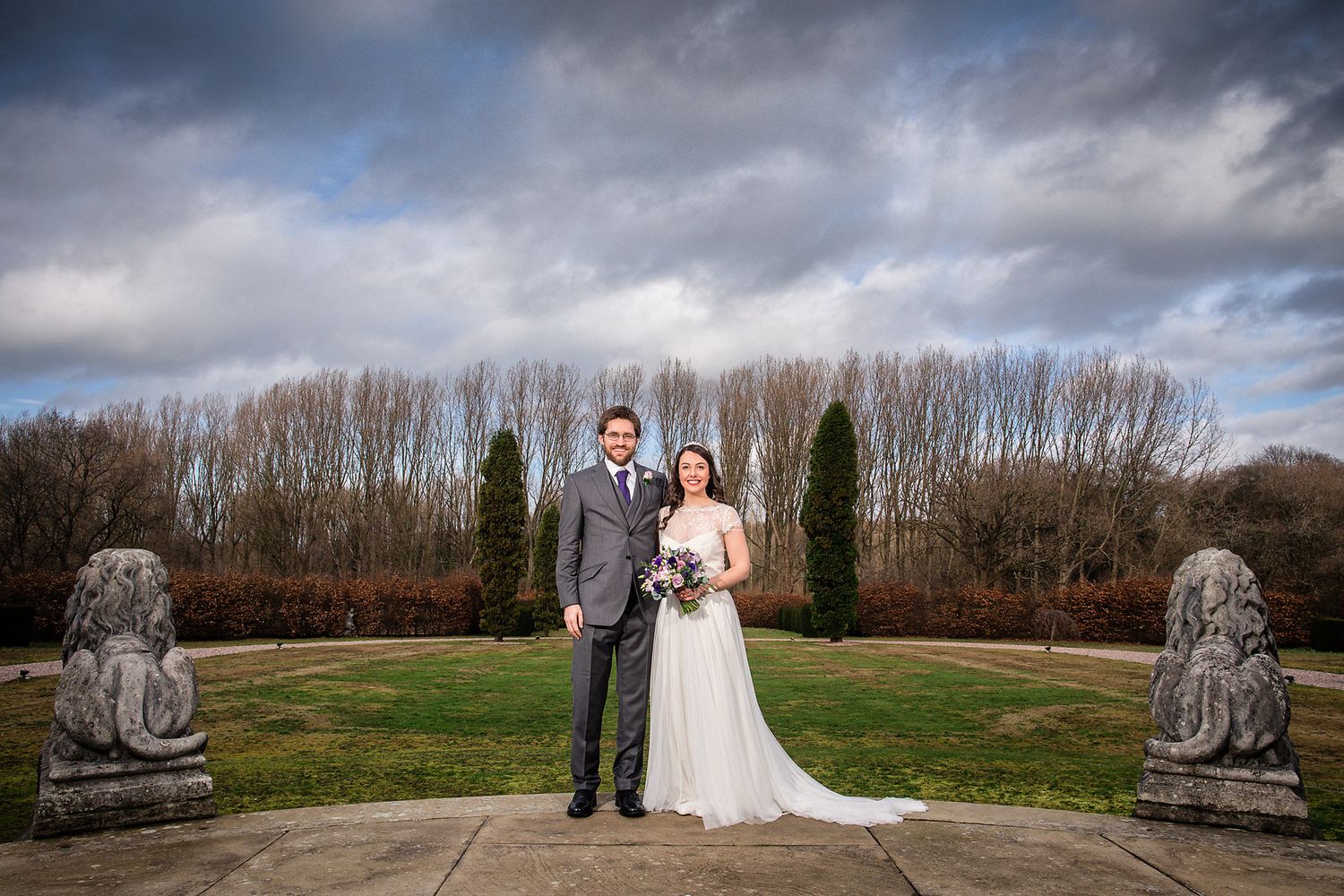 Wedding couple poses between stone lion statues in an elegant garden setting.