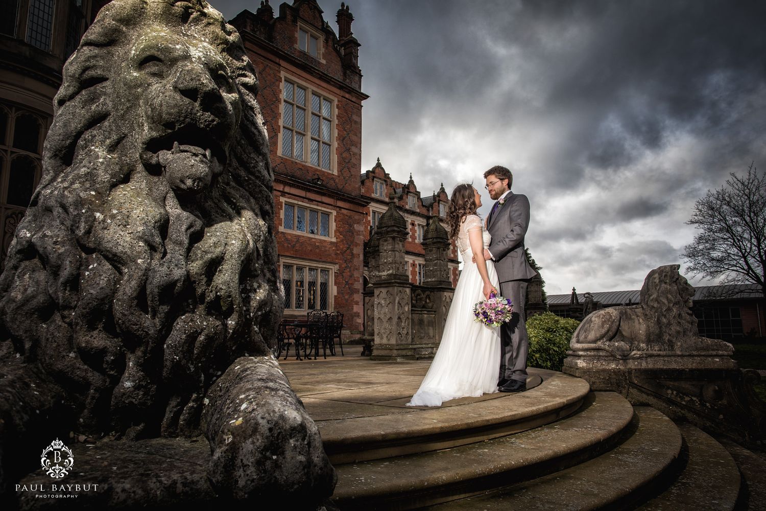 Newlyweds pose beside stone lion statue outside historic mansion with stormy sky.