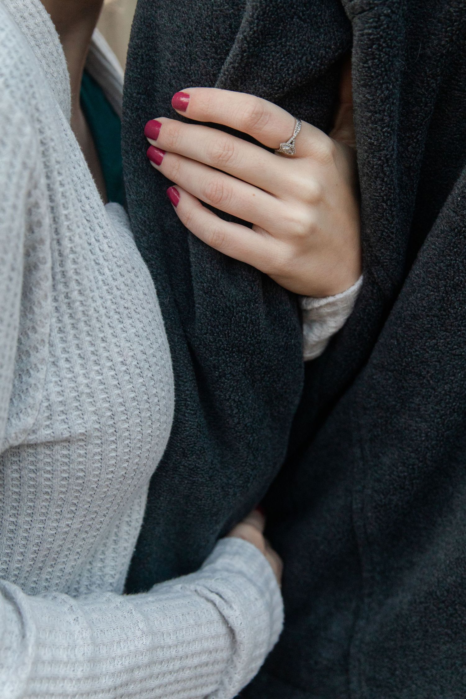 Close-up of a hand with burgundy nail polish and an engagement ring against a dark sweater.