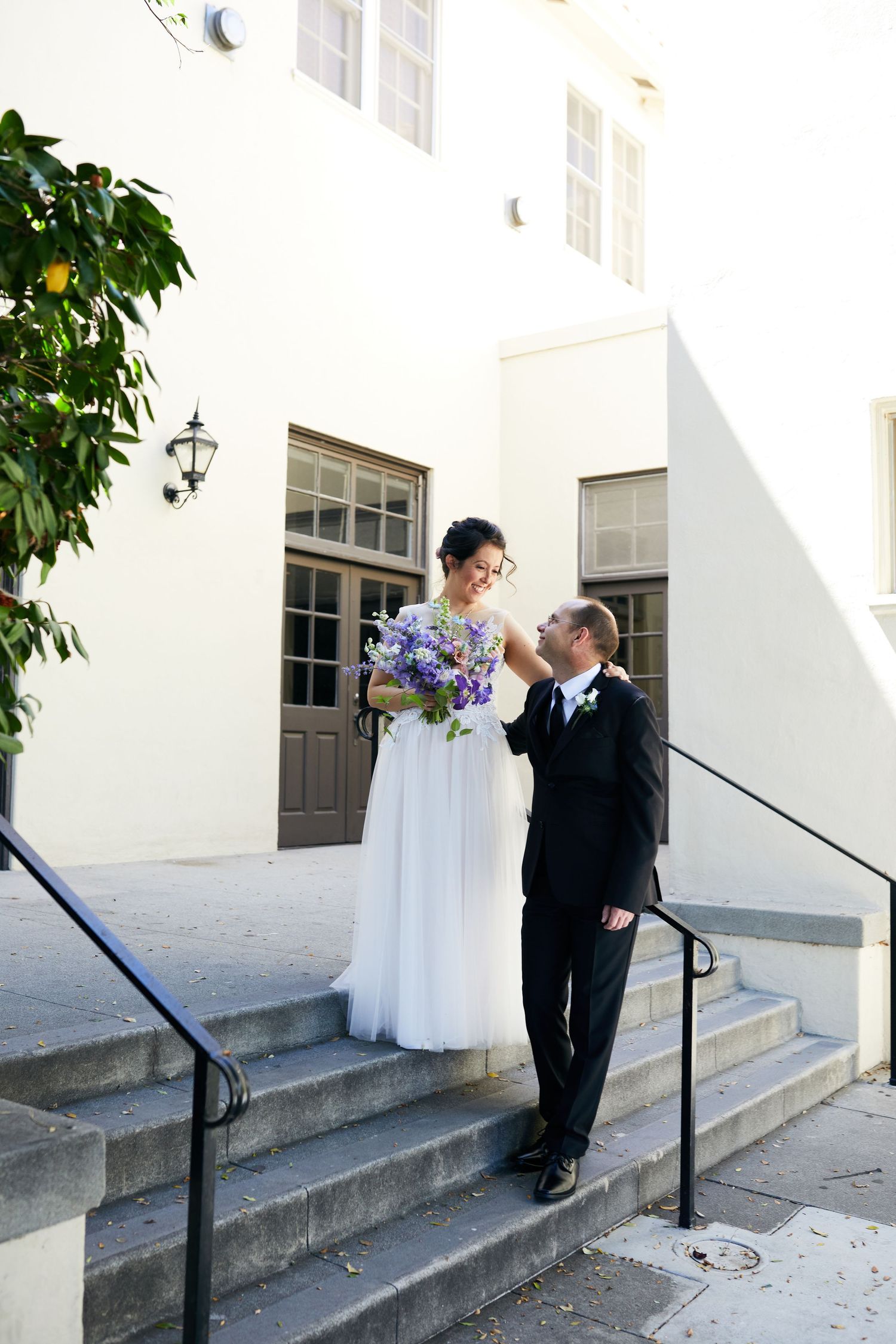 A couple stands on white church steps with purple flowers and elegant attire.