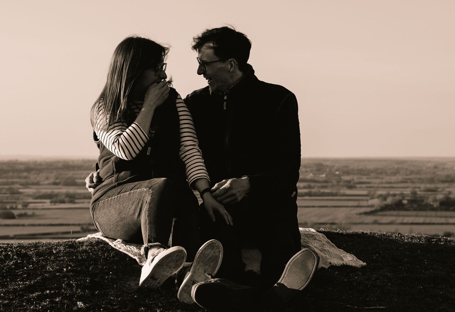 A black and white image of a young couple sat down with open views behind them. They are looking at each other and laughing.
