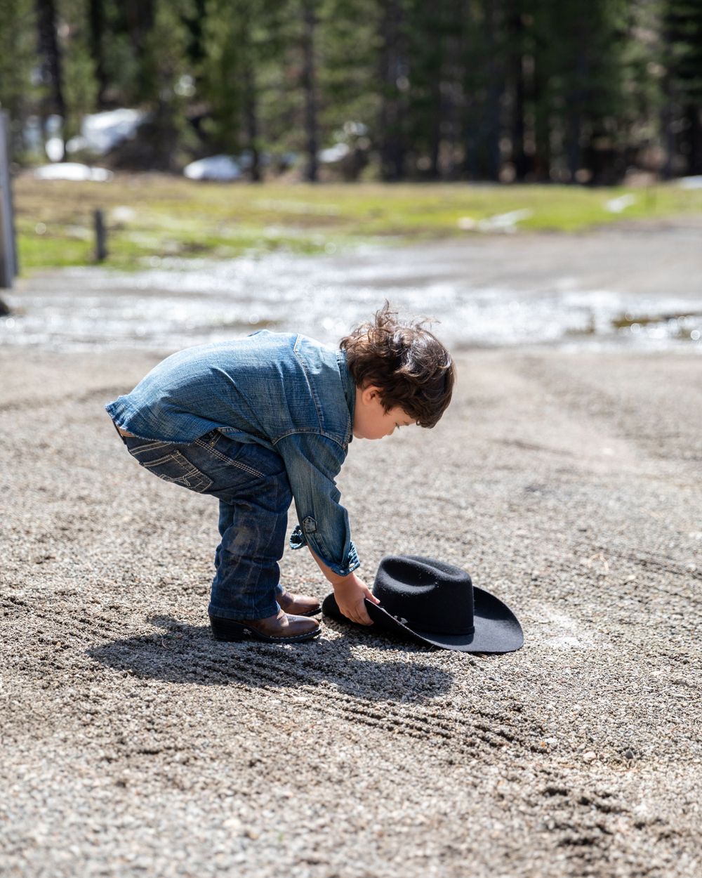 Orange County photographer captures Young child in denim outfit bends down to pick up a black cowboy hat from a gravel path.