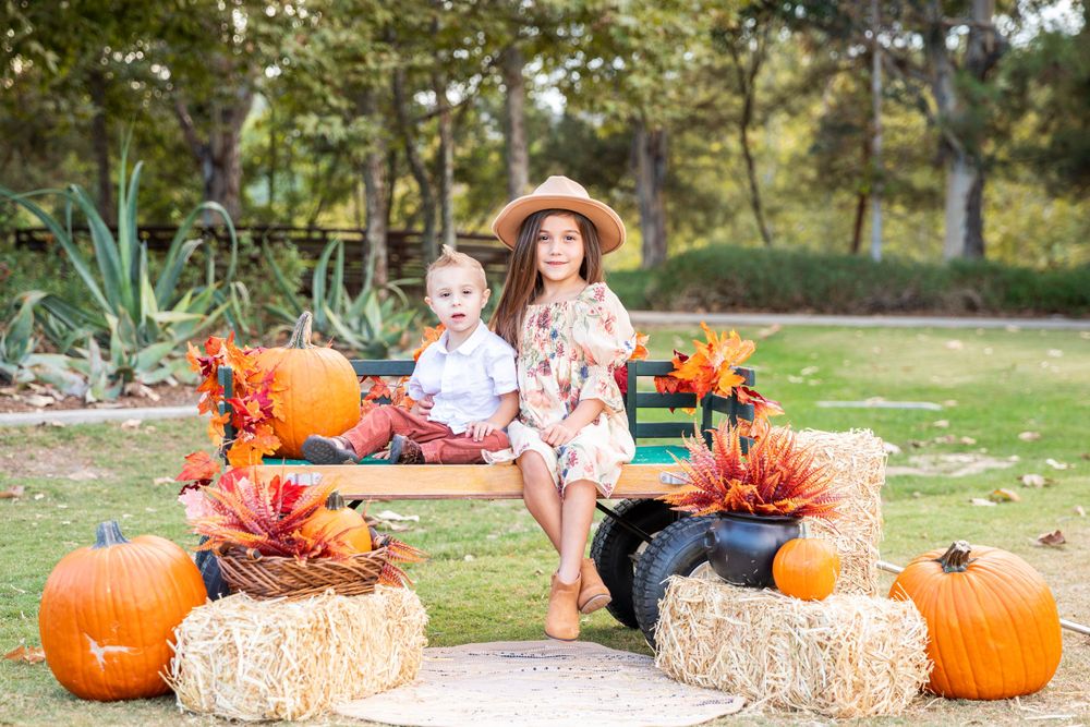 Orange County photographer captures Two children posing on bench surrounded by pumpkins and hay bales in fall outdoor portrait.