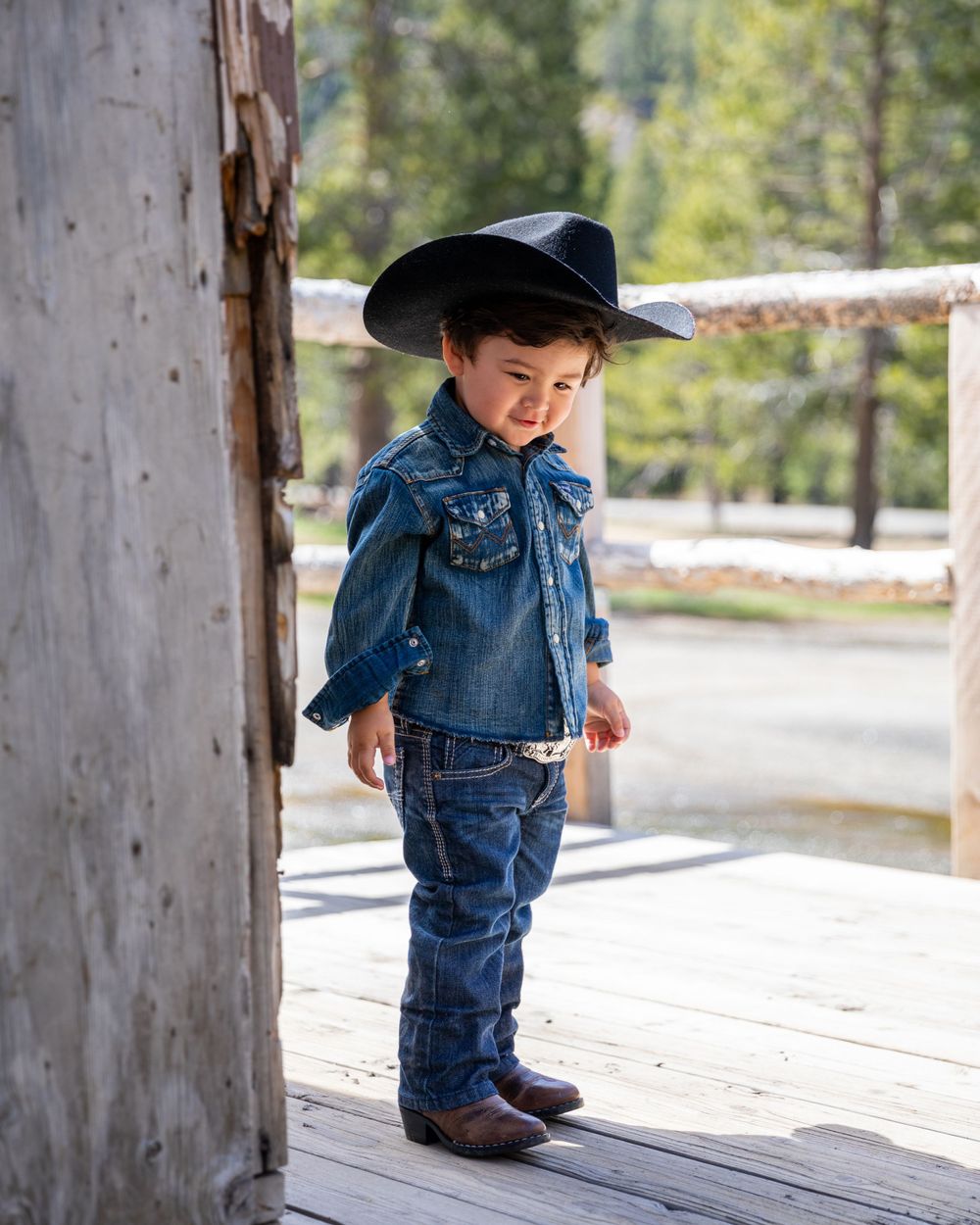 Orange County photographer captures Cute young boy in denim shirt and black cowboy hat stands on a wooden porch at a ranch.