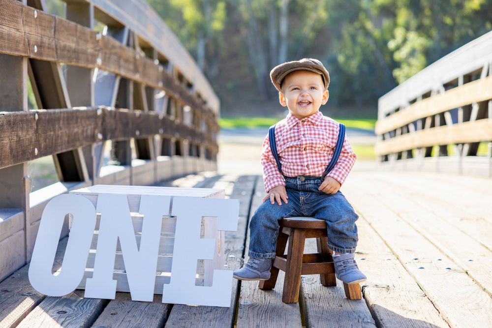 Orange County photographer captures Smiling baby boy in suspenders and flat cap sits on stool beside large ONE letters on a wooden bridge.