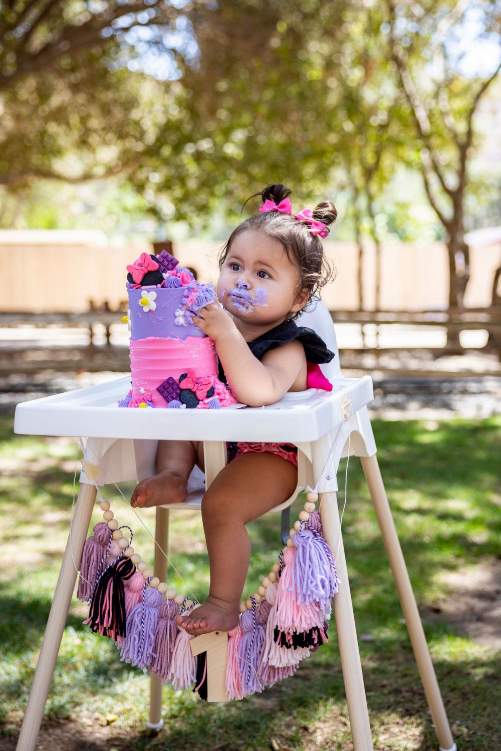 Orange County photographer captures girl with cake-smeared face hugging her birthday cake while sitting in a decorated high chair outdoors.