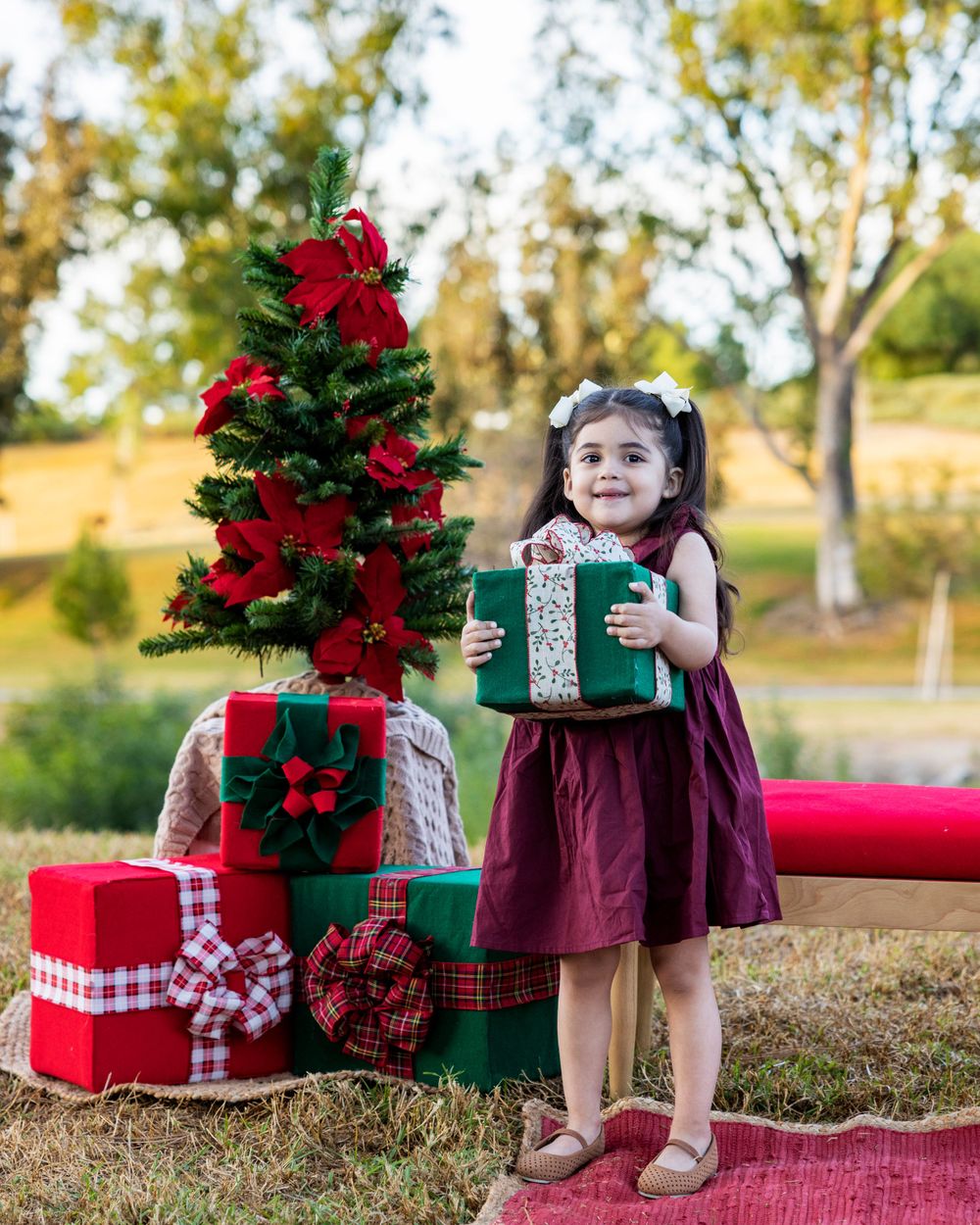 Orange County photographer captures little girl in burgundy dress holding a wrapped gift beside a decorated Christmas tree outdoors.