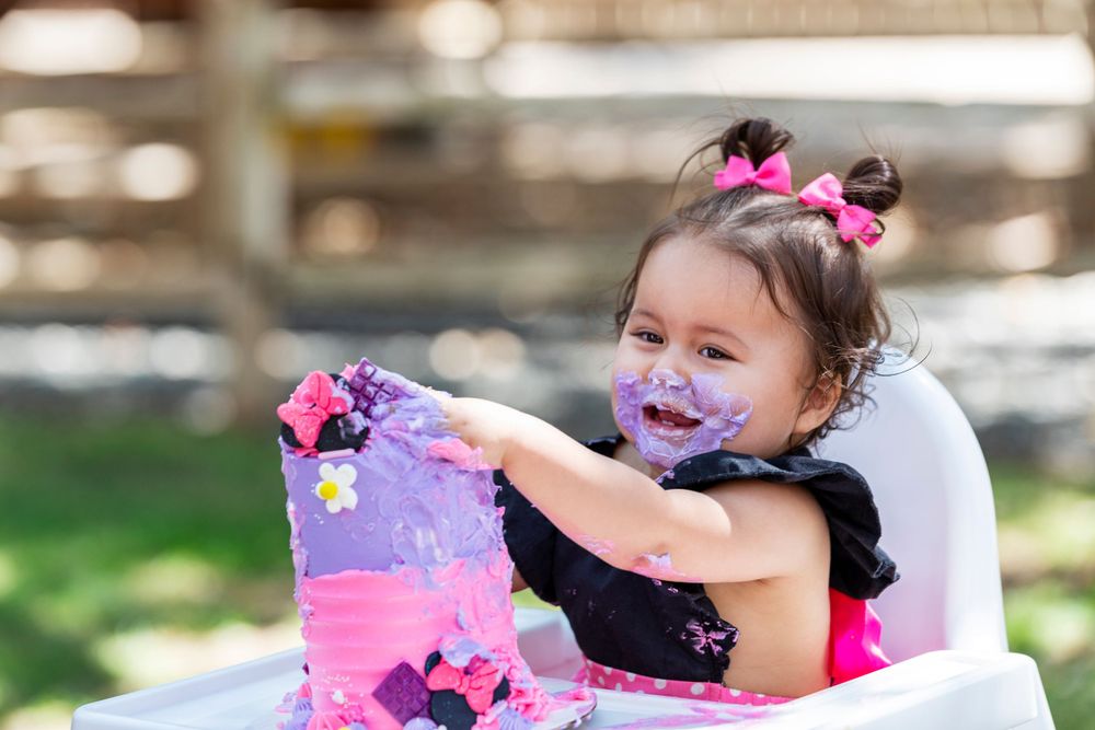 Orange County photographer captures toddler girl with frosting on her face enjoying a colorful cake smash celebration in a white high chair.