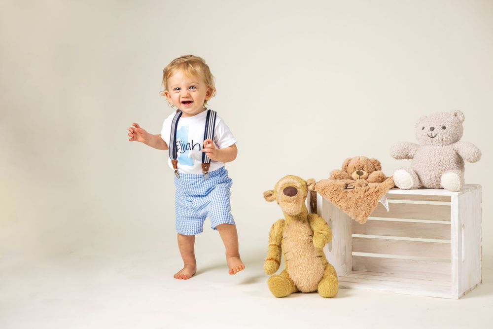 Orange County photographer Happy toddler boy in suspenders and blue shorts stands barefoot beside stuffed animal toys on white background.