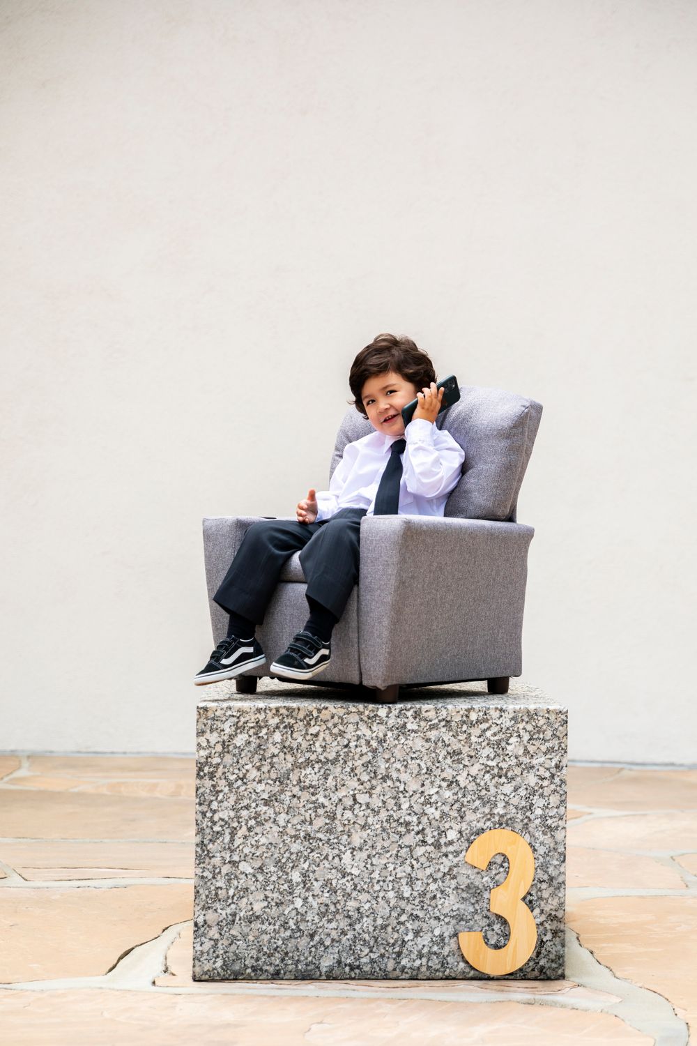 Orange County photographer Toddler boy in business suit talking on phone in gray armchair on stone pedestal with number 3.