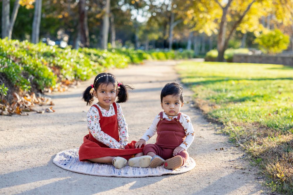 Orange County photographer captures siblings in matching red outfits sitting together on a path in a sunny autumn park setting.