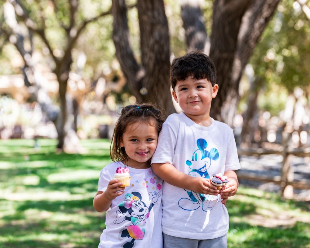 Orange County photographer captures smiling and holding ice cream cones together outdoors under shaded trees wearing Mickey Mouse shirts.