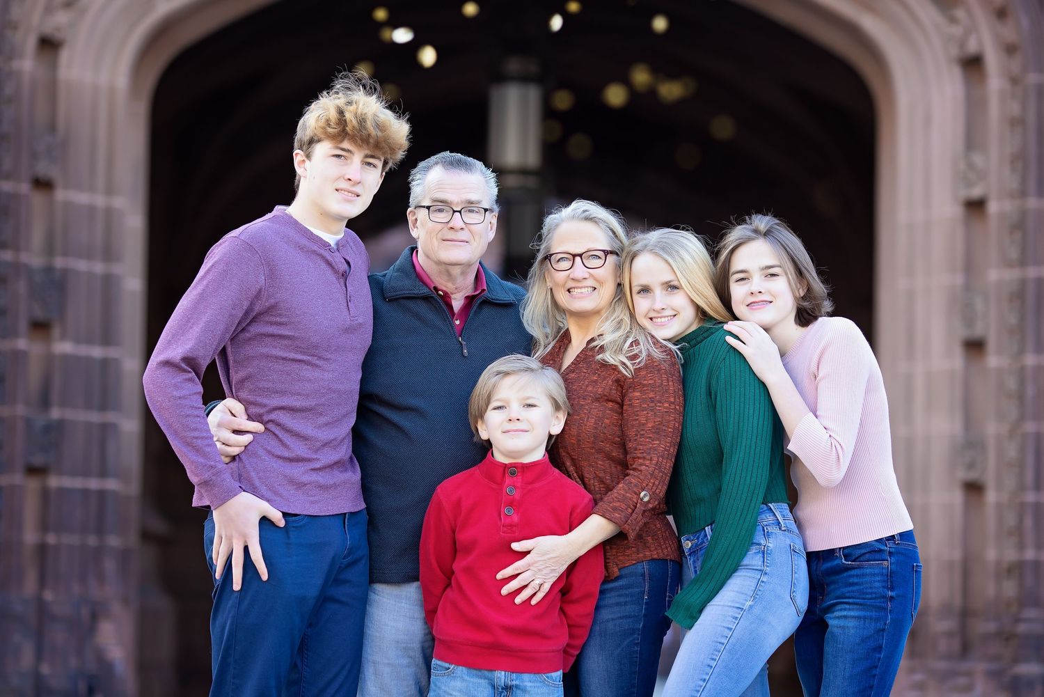 Family of six smiling together in front of a brick archway in an outdoor portrait.