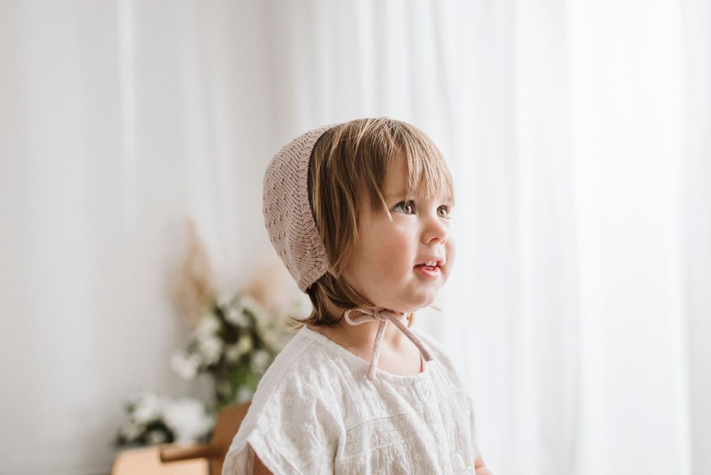 Young child in knit bonnet and white dress looking to side in bright interior.