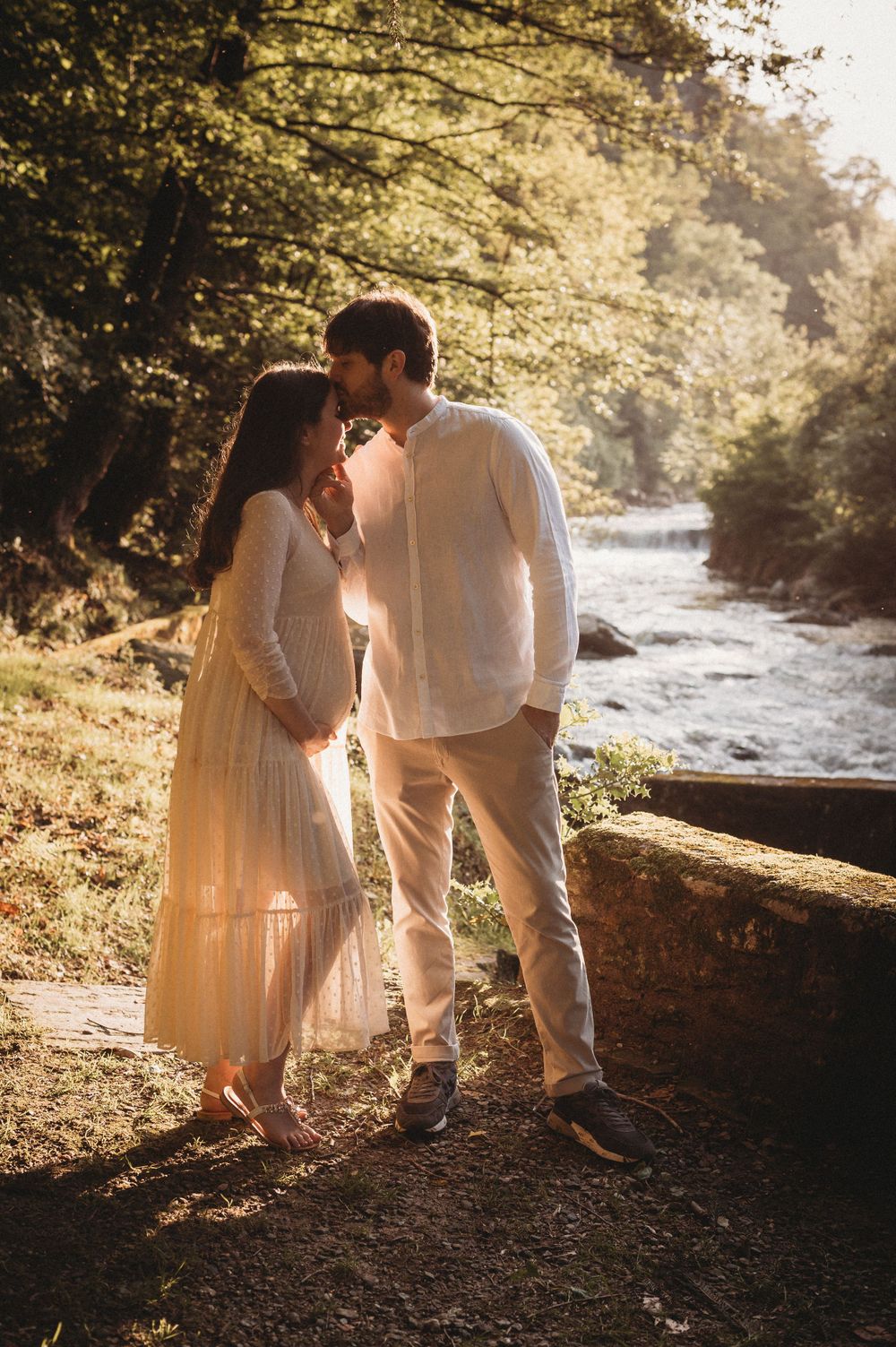 Romantic couple in light clothing embraces by river during golden sunset.