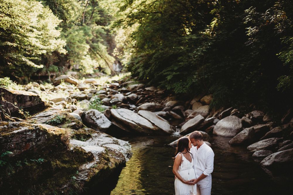 Romantic couple in white standing by rocky stream in lush forest setting.