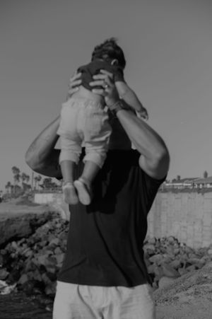 A black and white image of a playful moment with a child being lifted up against the sky.