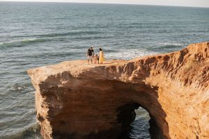 Two people stand atop a natural rock arch formation overlooking the ocean at sunset.