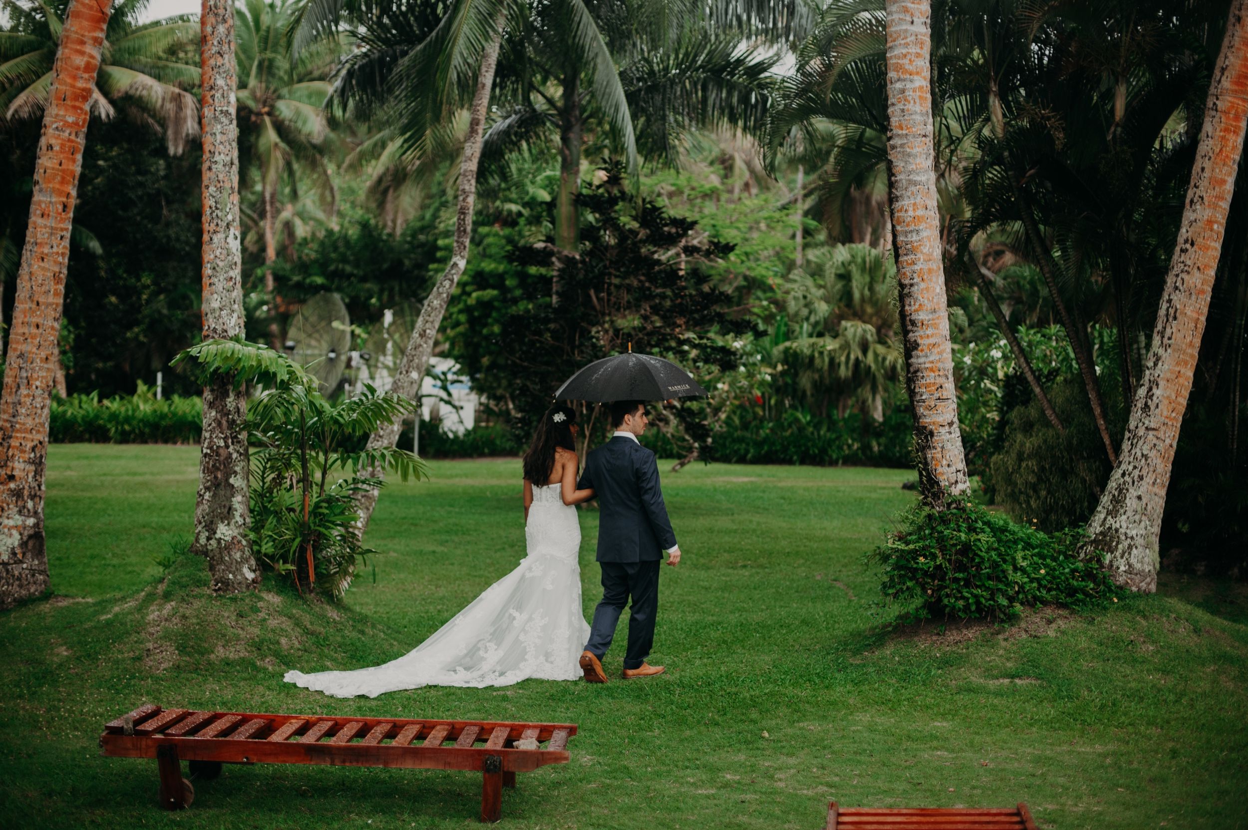 Wedding couple standing under palm trees in a lush tropical garden setting.