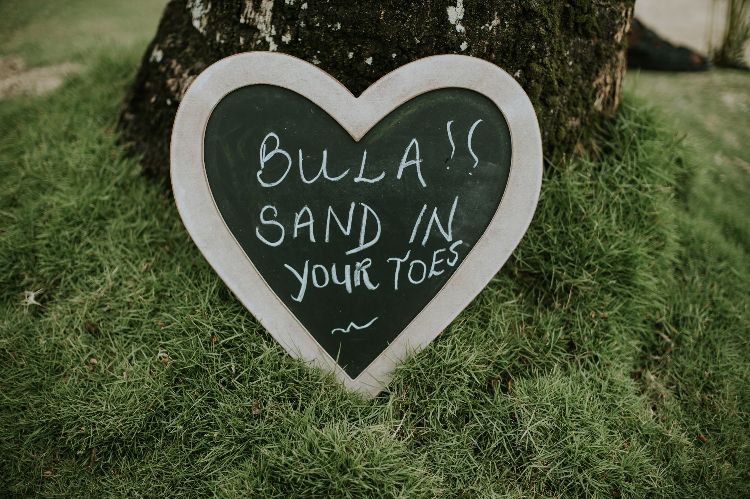 A heart-shaped chalkboard sign reading 'Bula sand in your toes' placed against green foliage.
