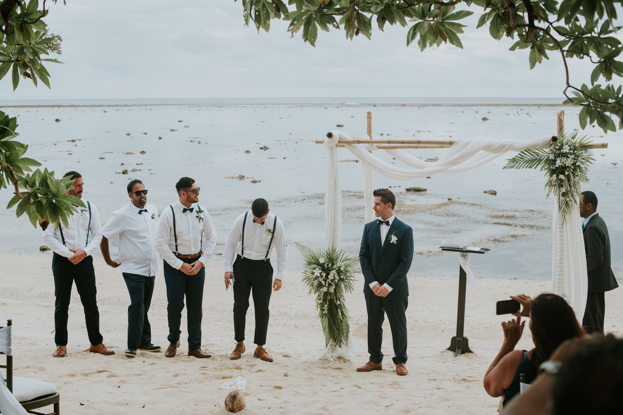 Beachfront wedding ceremony setup with wooden cross and groomsmen standing by the water.