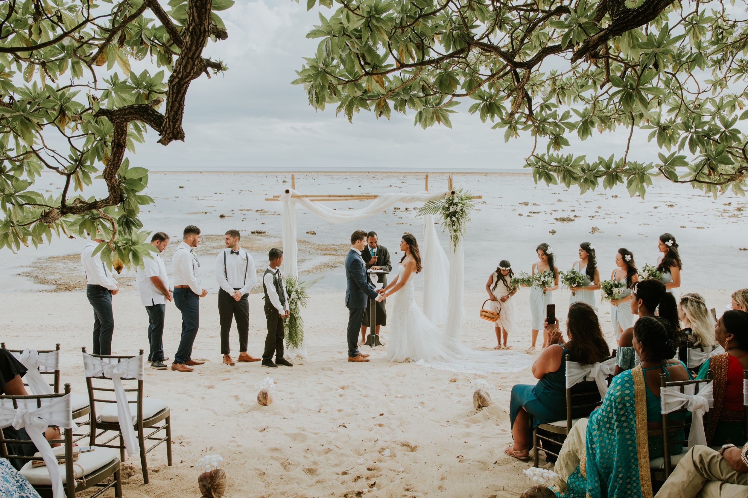 Guests gather on a beach for an outdoor wedding ceremony with palm trees and ocean views.