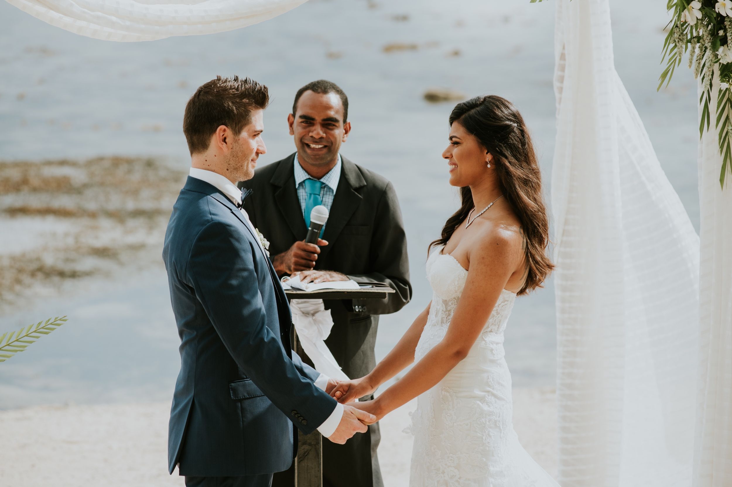 A couple exchanges vows during their beach wedding ceremony under a white arch with an officiant.