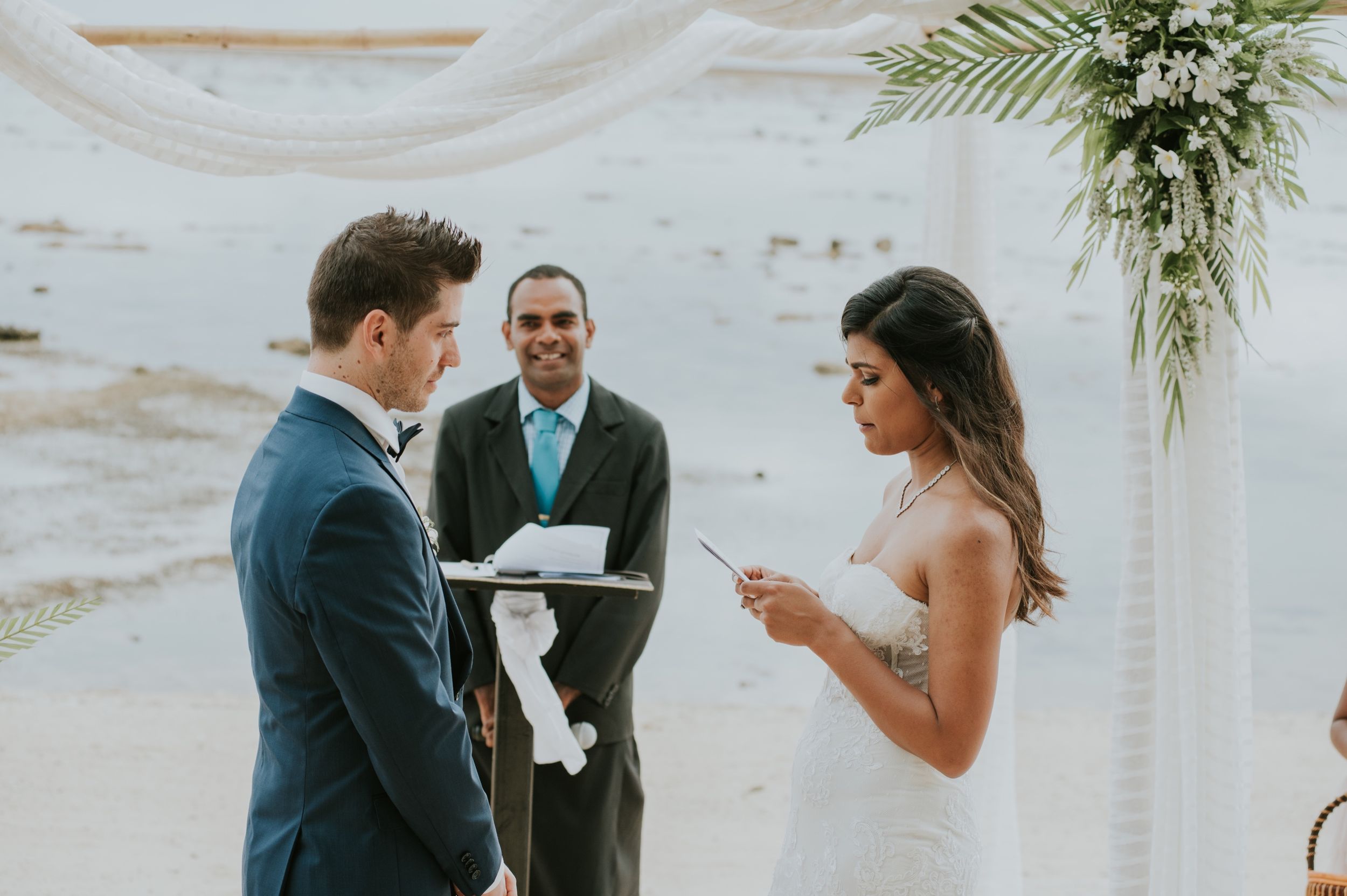 Wedding ceremony moment as couple exchanges rings under white arch with tropical decor.