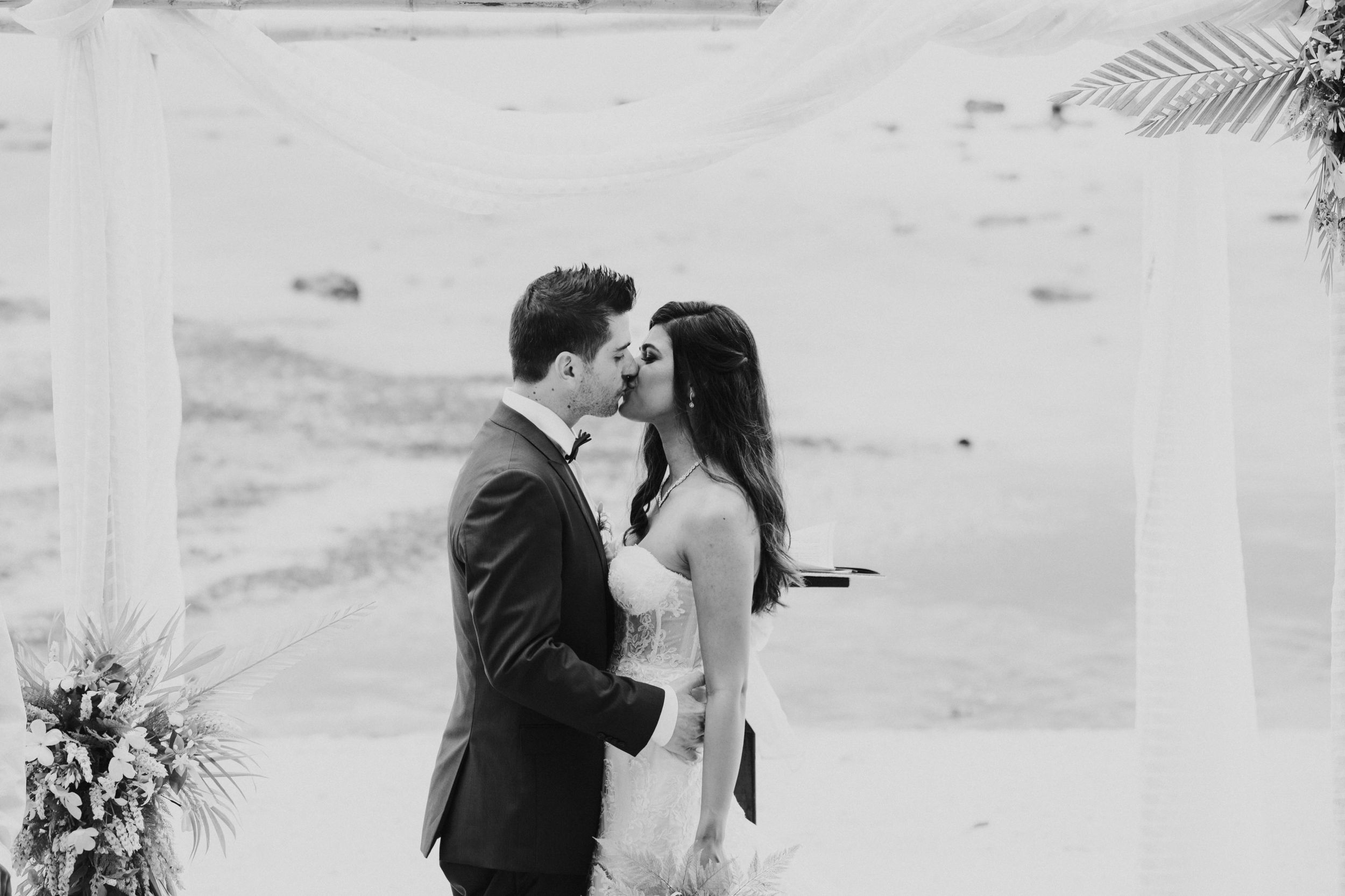 Black and white photo of newlyweds sharing their first kiss under white ceremony arch.