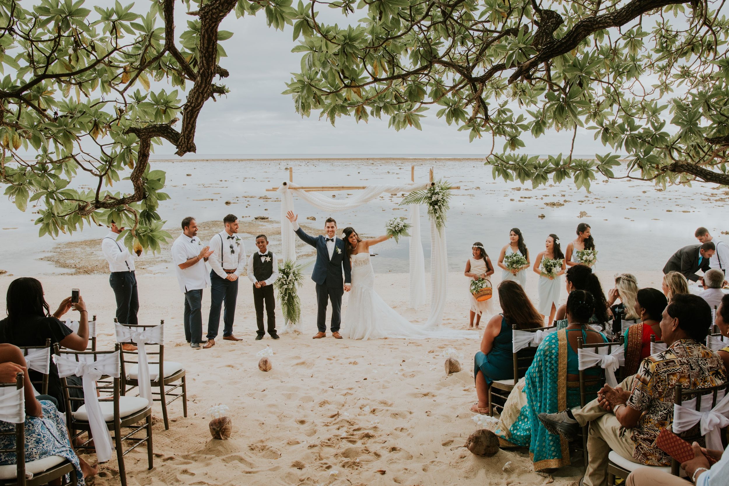 Beachside wedding ceremony with guests seated on both sides of sandy aisle under trees.