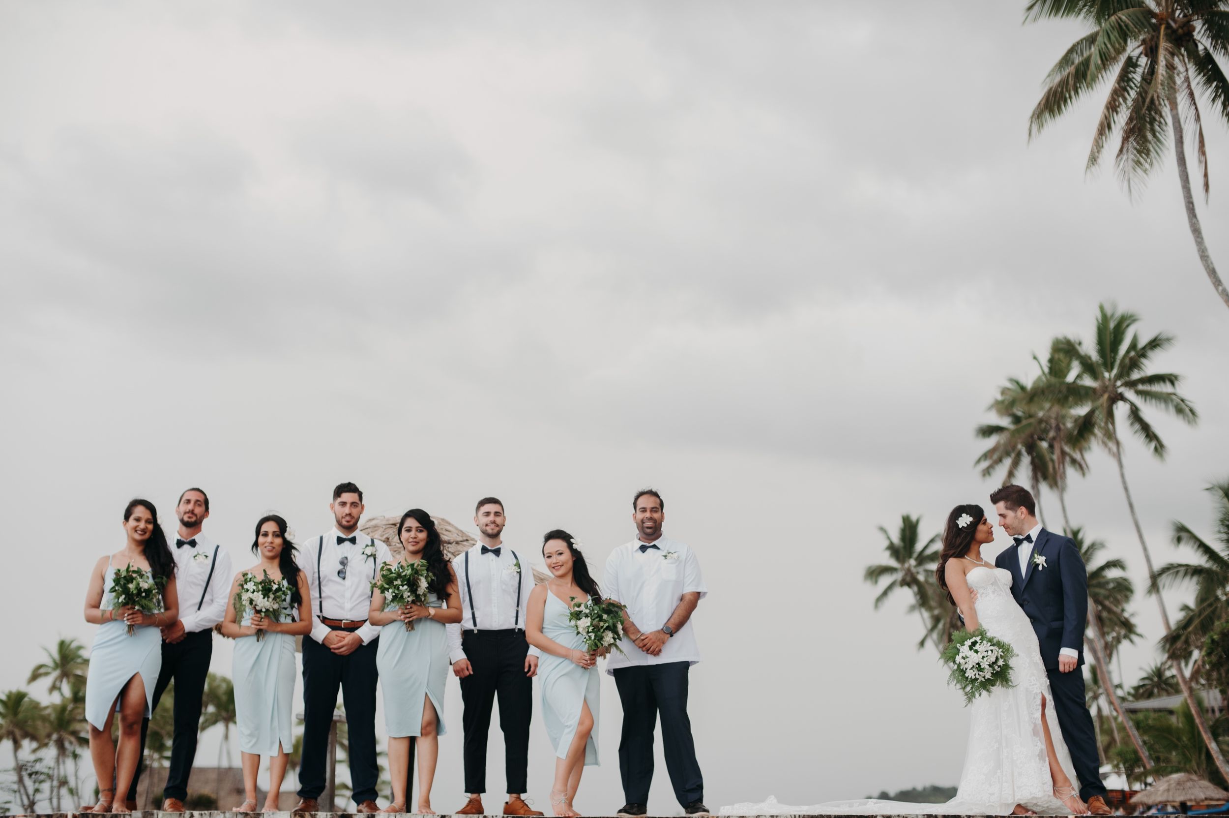 Wedding party standing together against palm trees on an overcast day.
