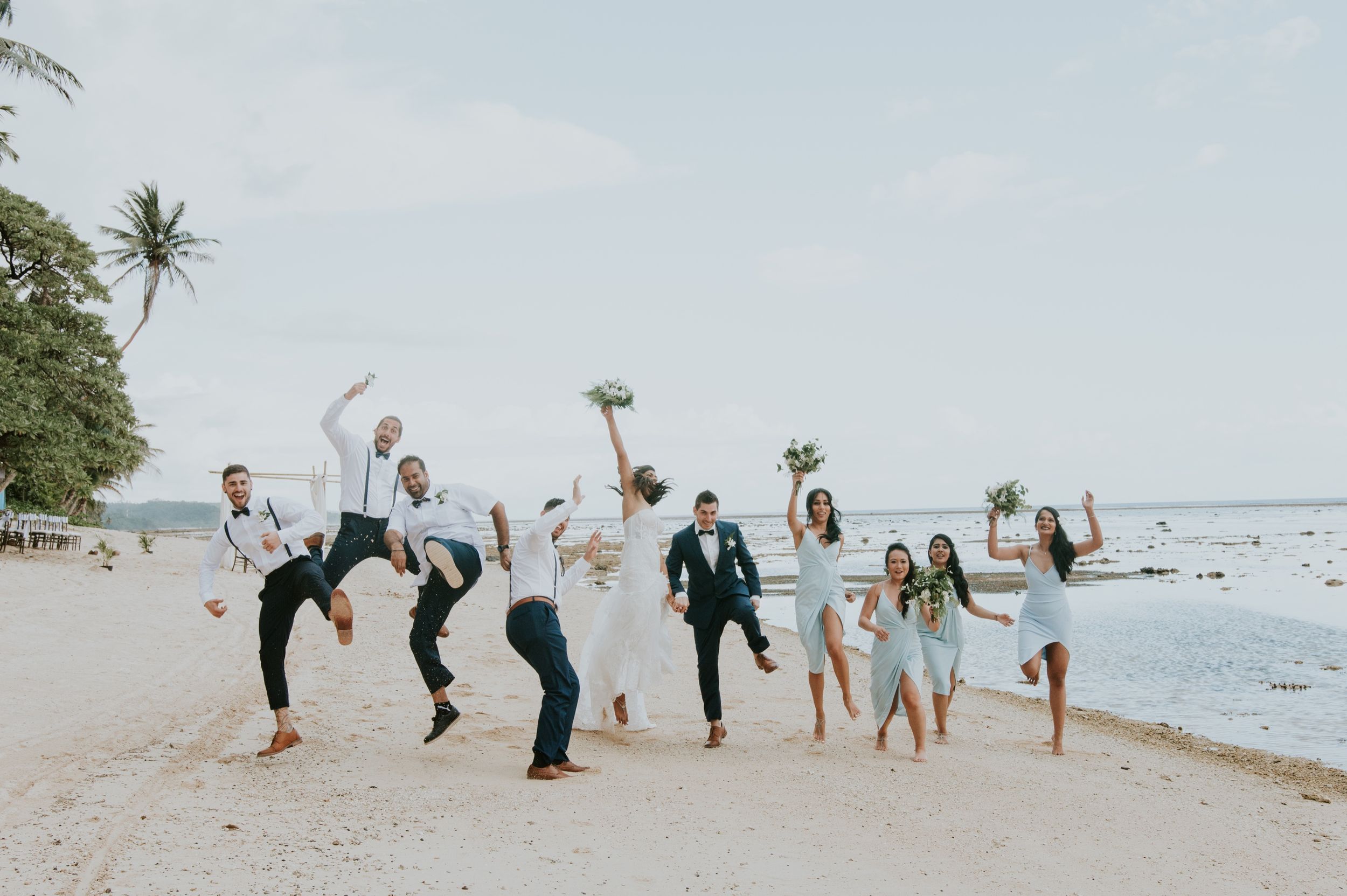 Wedding party jumping and celebrating together on a scenic beach with palm trees.