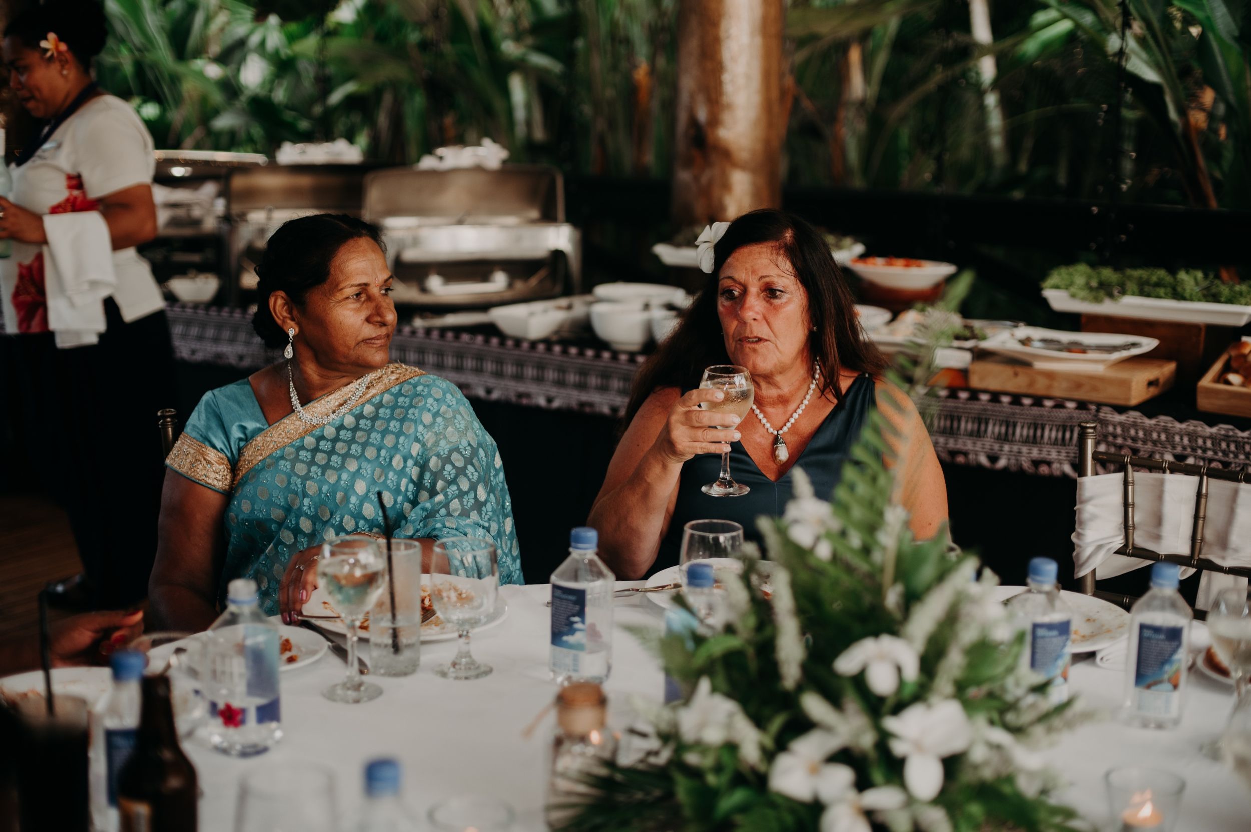 Wedding reception guests dining at decorated tables in tropical outdoor setting.