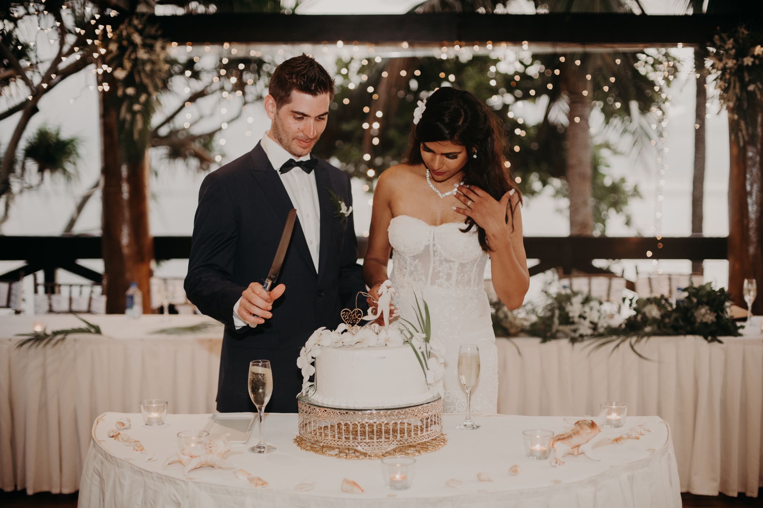 Newlyweds cutting their elegant white wedding cake together at an outdoor reception with twinkle lights.
