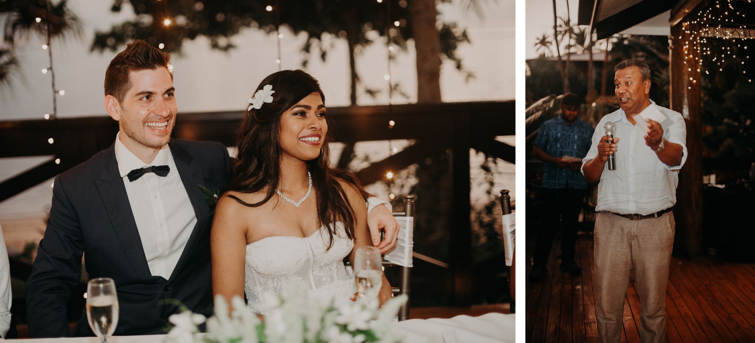 Bride and groom listening to toasts at reception head table while guest speaks in background.