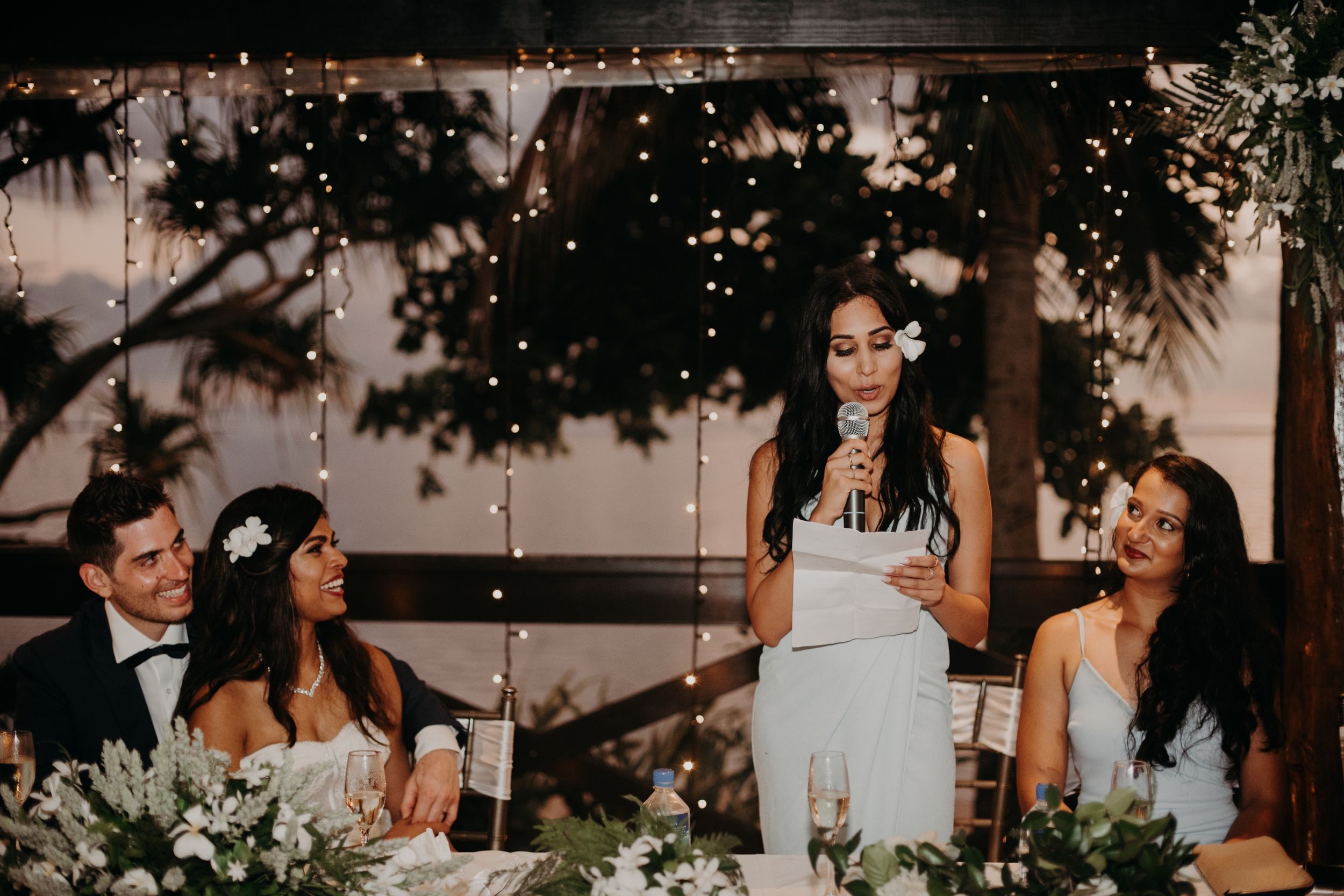 Wedding party reacting to speeches at decorated head table during evening reception.